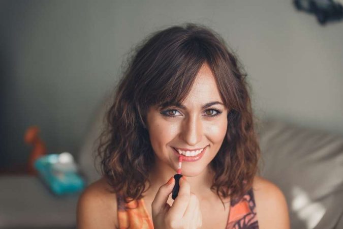 Une femme aux cheveux bruns mi-longs onduleux sourit en appliquant du rouge à lèvres liquide rose corail sur sa bouche avec un petit applicateur.